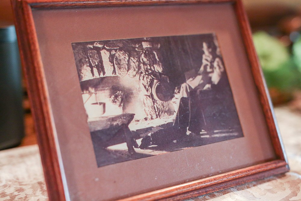 A sepia-toned photograph in a wooden frame shows a person sitting by a fireplace, surrounded by hanging pots and rustic decor, creating a cozy, vintage atmosphere that echoes the rich Gold Mountain Manor history.