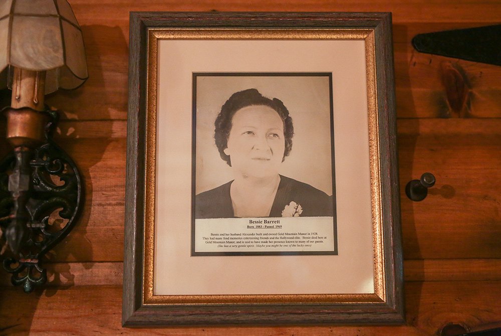 Framed black-and-white portrait of a woman with short dark hair, wearing a dark top and a brooch, displayed on a wooden wall, next to a vintage lamp and small decorative items—a striking vignette that captures the timeless elegance woven throughout Gold Mountain Manor history.
