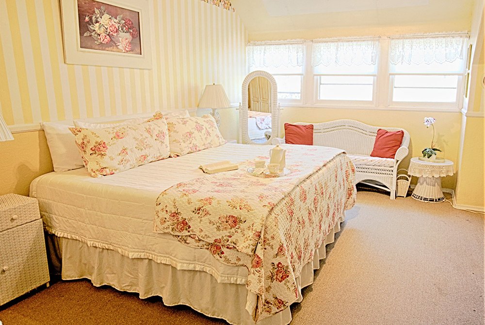 Bright bedroom with a king bed, floral bedding, wicker furniture, a mirror, lace curtains, and a small table with a flower. The decor features soft yellow walls and a cozy, vintage style reminiscent of charming Big Bear bed and breakfast rooms.