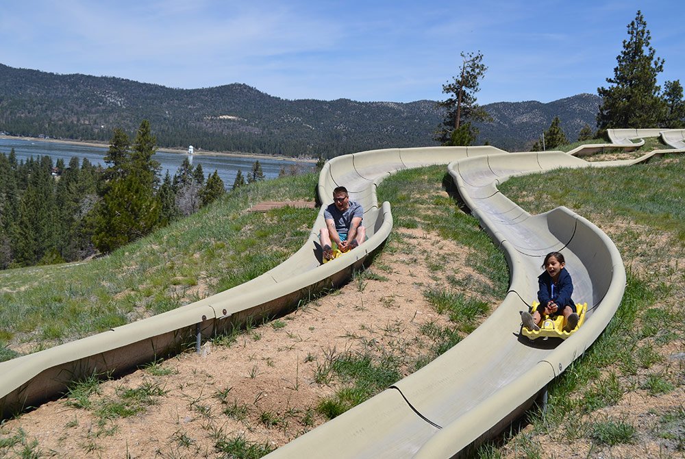 Two people ride separate concrete alpine slides down a grassy hillside with trees, alongside a lake and surrounded by forested mountains under a clear blue sky—one of the exciting things to do in Big Bear Lake.
