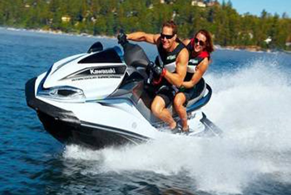 Two people wearing life jackets ride a white and black Kawasaki jet ski at high speed on a lake, with water splashing behind them and trees visible in the background—a thrilling example of the exciting things to do in Big Bear Lake.