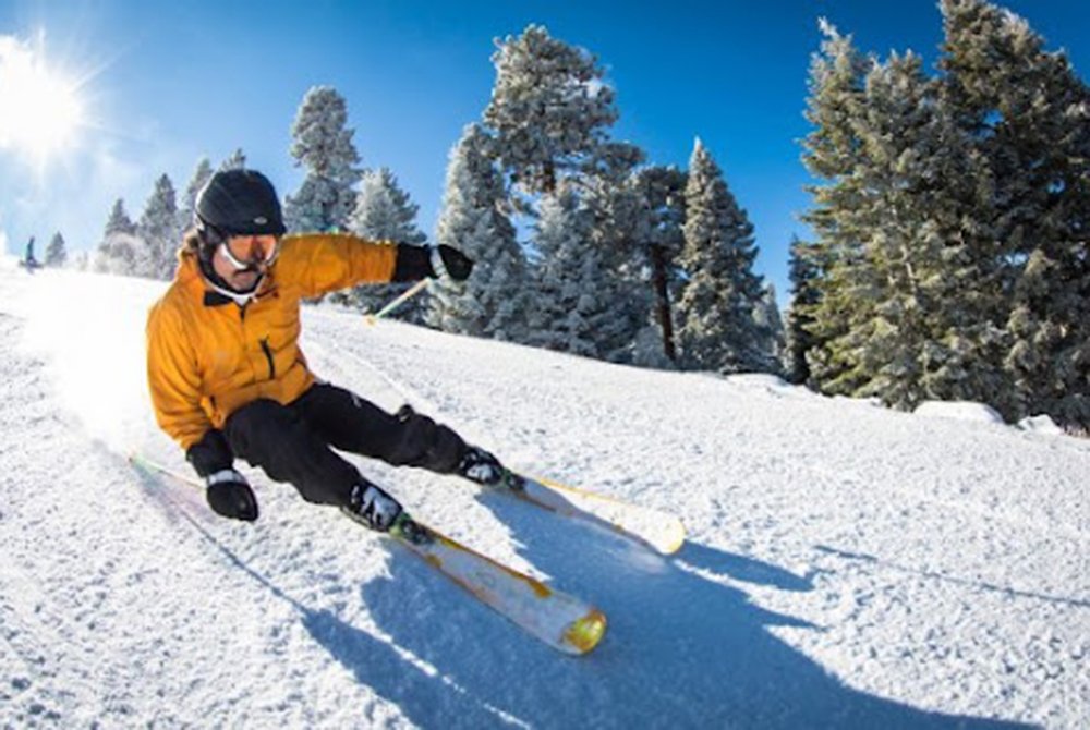 A skier in a yellow jacket and helmet swiftly carves down a snowy slope with pine trees and a bright blue sky in the background—a thrilling highlight among the many things to do in Big Bear Lake.