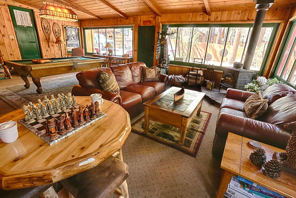 Cozy wooden cabin interior with leather couches, a coffee table, wood stove, pool table, and a small round table with a chess set. Large windows let in natural light, creating the perfect retreat for guests seeking a classic Big Bear bed and breakfast experience.
