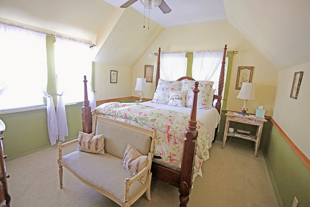 Cozy bedroom with a four-poster bed, floral bedding, two nightstands with lamps, a small upholstered bench at the foot of the bed, and large windows with sheer curtains letting in natural light—just the kind of inviting space you might find in charming Big Bear bed and breakfast rooms.