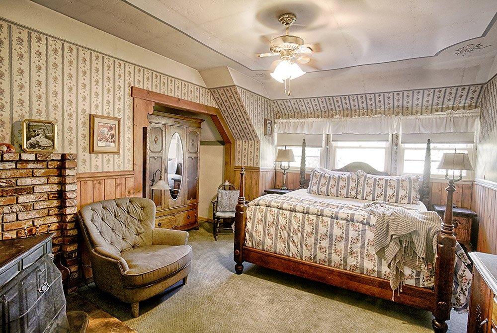 A cozy vintage bedroom reminiscent of charming Big Bear bed and breakfast rooms, with floral wallpaper, a wooden bed, a cushioned chair, an armoire, a brick fireplace, and windows letting in natural light. A ceiling fan and lamps add warmth to the space.