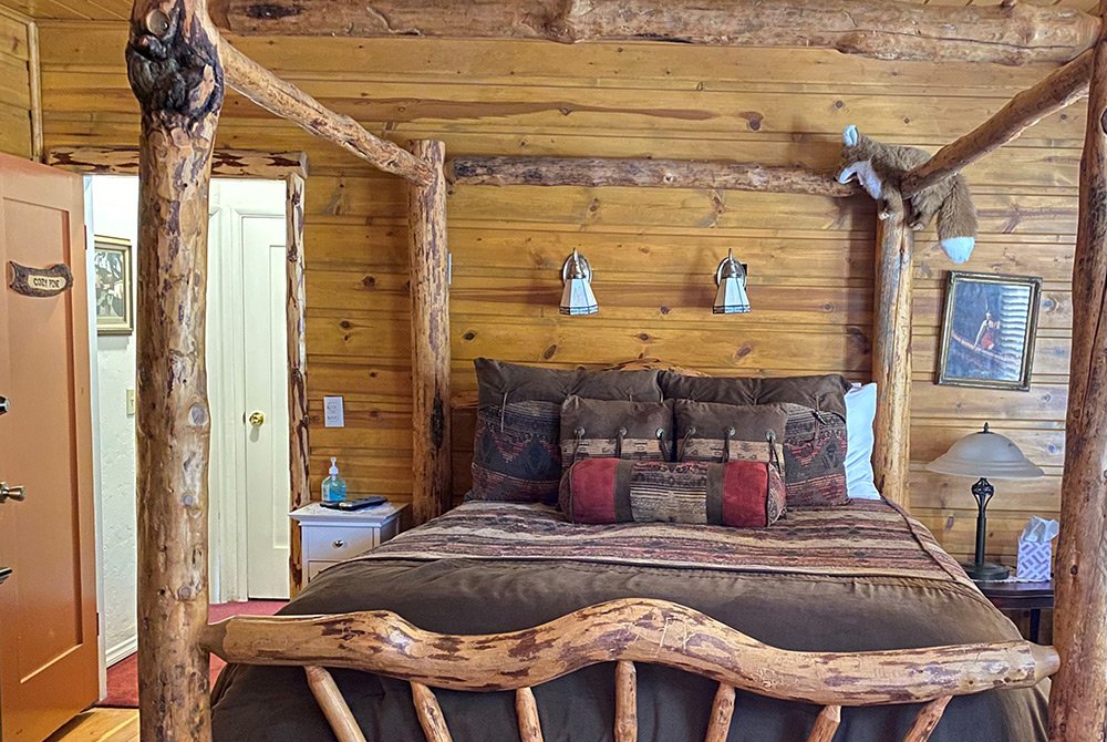 Rustic log cabin bedroom with a wooden four-poster bed, earth-toned bedding, wall-mounted lamps, and animal decor. Wooden walls and door add a cozy, natural feel reminiscent of classic Big Bear bed and breakfast rooms. A small table with a lamp sits beside the bed.