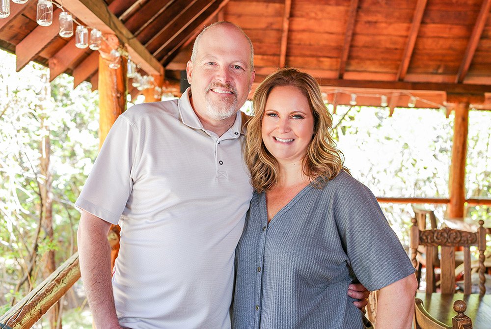 A man and woman stand close together, smiling at the camera on a wooden porch with a rustic roof and greenery in the background, capturing a warm moment at a historic Big Bear bed and breakfast.