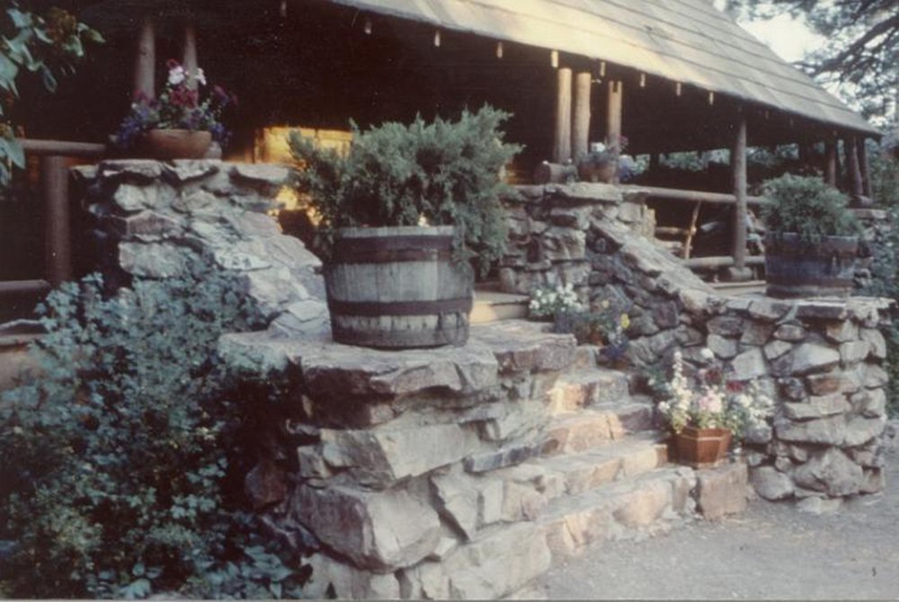 A rustic stone staircase leads to a wooden porch, with potted plants and barrels on the steps and near the entrance—a charming nod to Gold Mountain Manor history. The porch features wooden railings and is partially shaded by a roof, evoking the timeless character of this historic retreat.