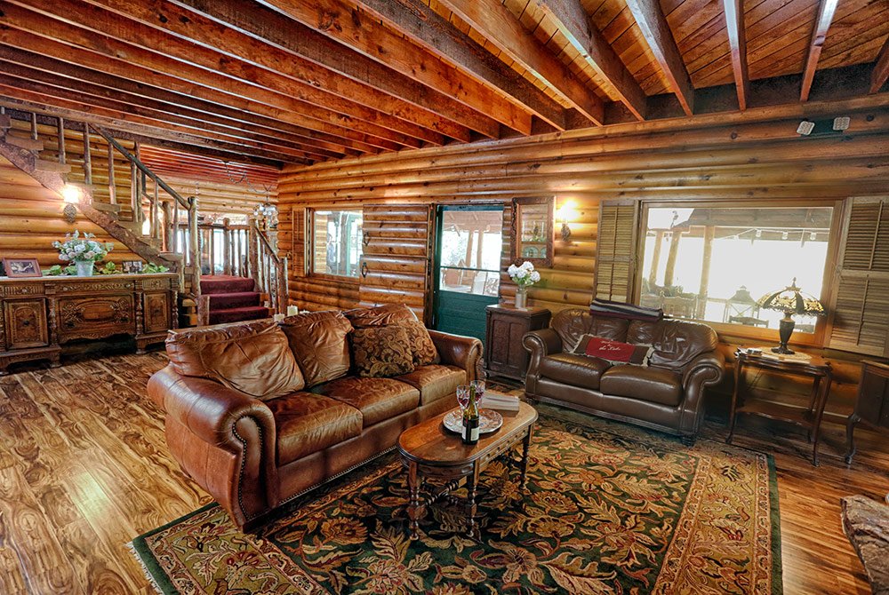 Cozy log cabin living room with exposed wood beams, brown leather sofas, a patterned rug, and wooden furniture. Perfect for Big Bear group events, this inviting space features stairs leading up in the background and windows that let in plenty of natural light.