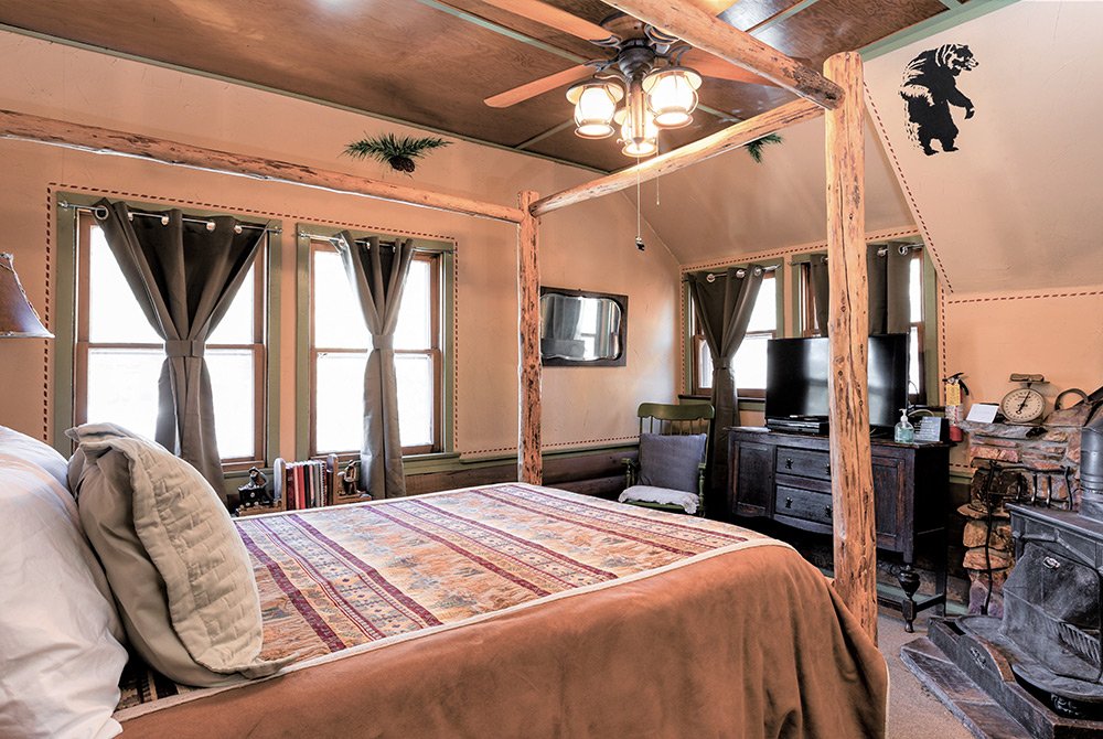 Cozy rustic bedroom with a wooden four-poster bed, patterned bedding, multiple windows with curtains, a television, fireplace, and bear wall art. Warm lighting enhances the inviting atmosphere—just like Big Bear bed and breakfast rooms known for their charm and comfort.
