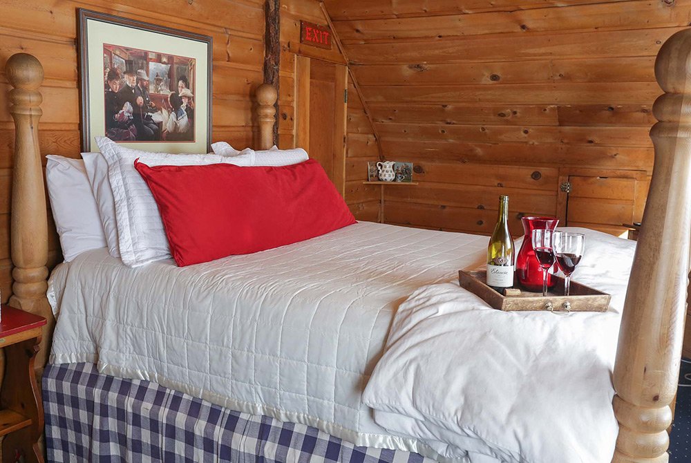 A cozy wooden bedroom with a neatly made bed, red and white pillows, and a tray holding wine, glasses, and a decanter. A painting hangs above the bed, capturing the inviting charm of classic Big Bear bed and breakfast rooms, while the room exudes a rustic, cabin-like feel.