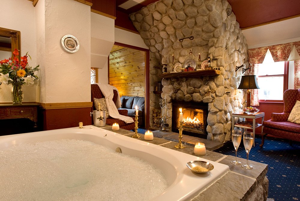 A cozy room at a Big Bear bed and breakfast features a bubbling hot tub near a stone fireplace, lit candles, champagne glasses, and elegant furniture, creating a warm, inviting atmosphere.