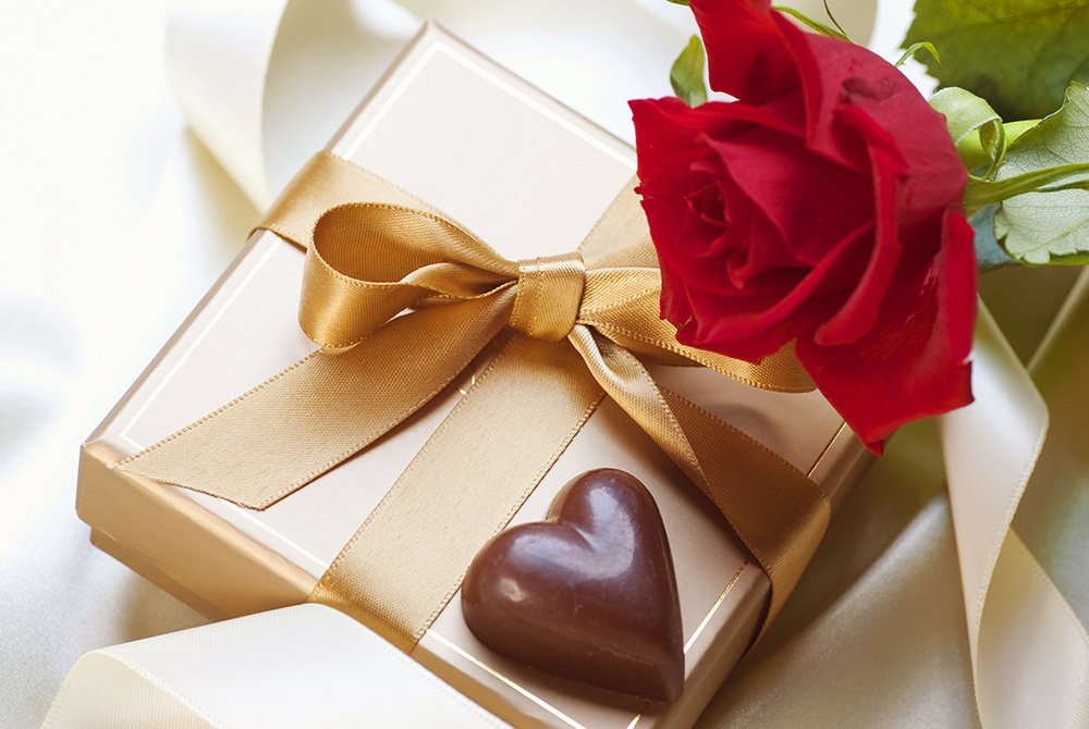 A gold gift box with a matching ribbon, a red rose, and a heart-shaped chocolate are elegantly arranged on a white satin surface—perfect touches for an unforgettable Big Bear romance package.