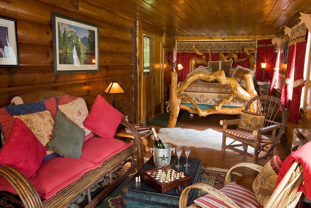 Cozy log cabin room with wooden furniture, a rustic bed, colorful pillows, a chessboard, and champagne on a table. Warm lighting and nature-themed decor create a comfortable and inviting atmosphere reminiscent of charming Big Bear bed and breakfast rooms.