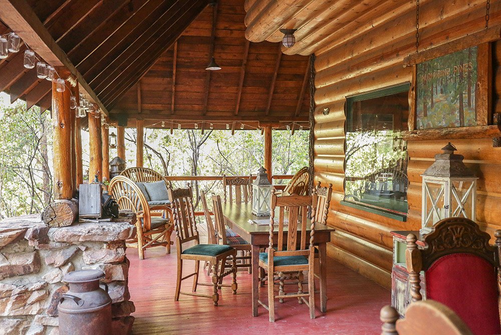 A rustic wooden porch at a charming Big Bear bed and breakfast features a long dining table, wooden chairs, wicker seating, lanterns, and a stone wall, all surrounded by trees and lush greenery.