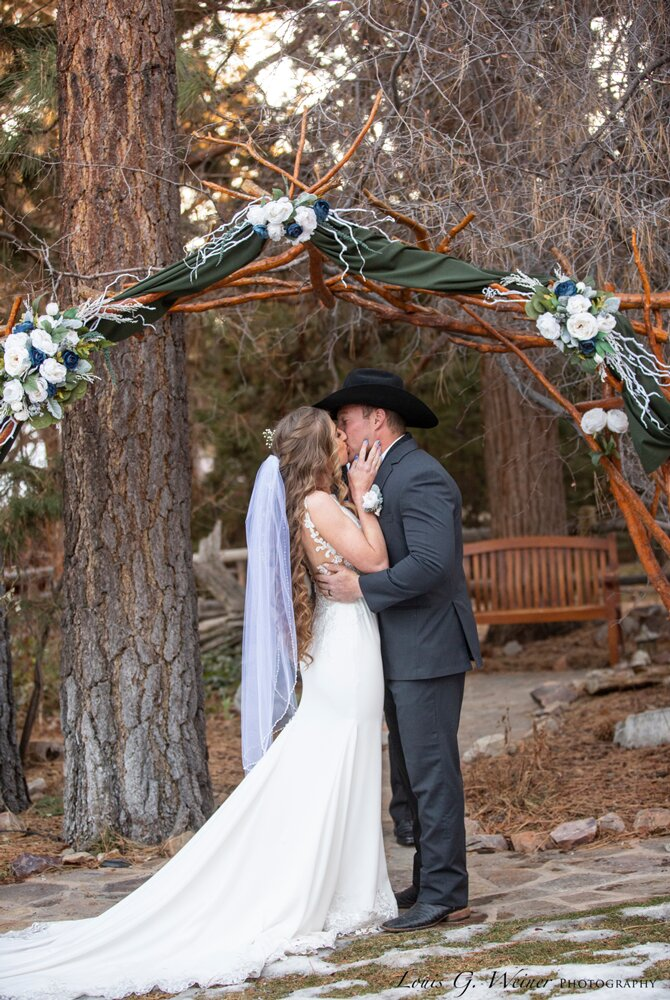 A bride and groom kiss under a rustic, flower-decorated arch outdoors, surrounded by trees in the heart of Big Bear wedding seasons. The bride wears a white dress and veil; the groom wears a dark suit and cowboy hat.
