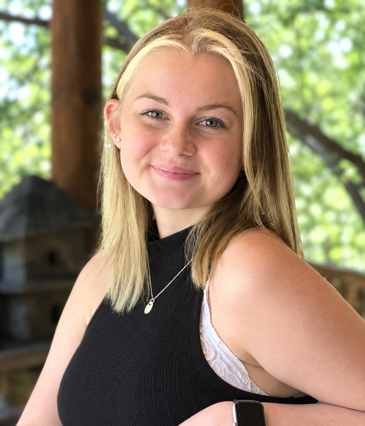 A young woman with blonde hair and a black sleeveless top smiles slightly, standing outdoors with trees and sunlight in the background, capturing the relaxed charm of a historic Big Bear bed and breakfast.
