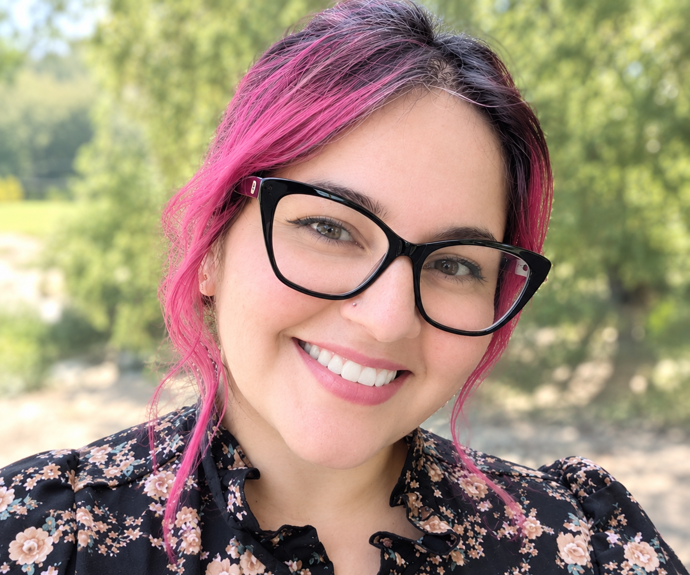Woman with pink hair and black glasses smiles outdoors, wearing a black floral shirt. Green trees and sunlight create a soft, natural background, reminiscent of relaxing mornings at a historic Big Bear bed and breakfast.