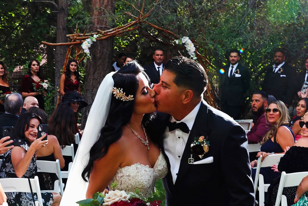 A bride and groom kiss at their outdoor wedding ceremony, surrounded by guests seated on white chairs, with a natural wooden arch and lush greenery in the background—a perfect scene inspired by the romantic atmosphere of Big Bear wedding seasons.