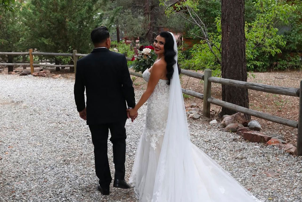 A bride in a white gown and groom in a black suit walk hand-in-hand outdoors on a gravel path, surrounded by trees. Capturing the magic of Big Bear wedding seasons, the bride looks back and smiles, holding a bouquet of flowers.