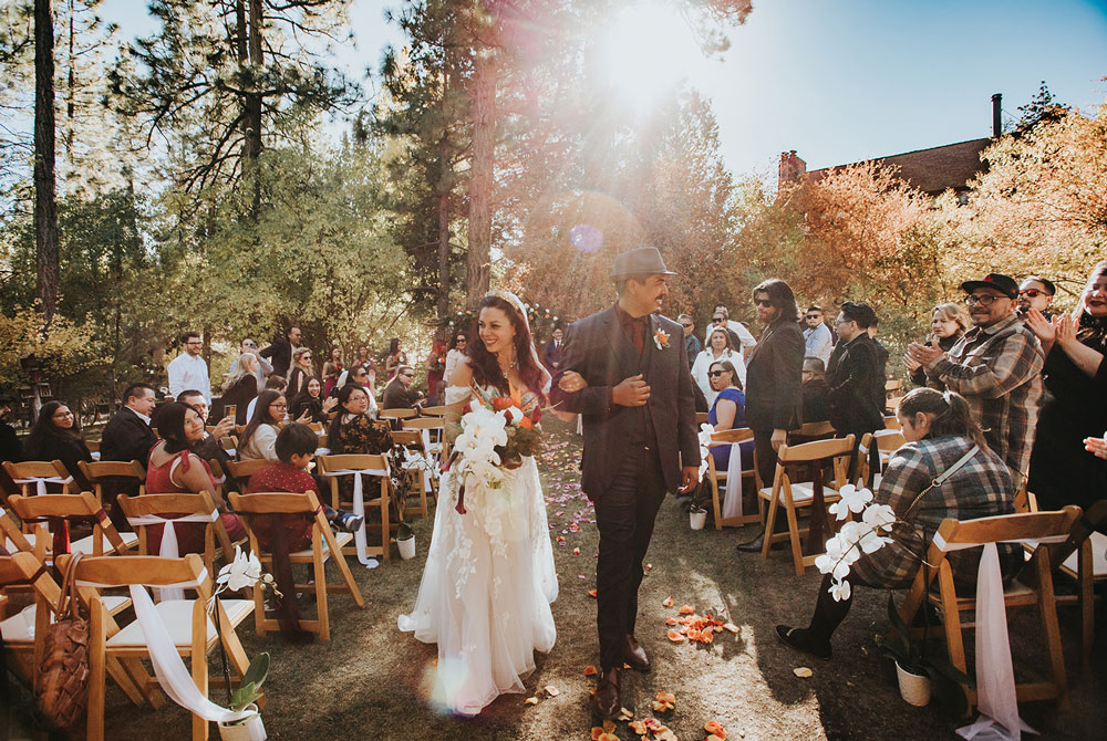 A bride and groom walk down the outdoor aisle smiling, surrounded by guests seated on wooden chairs and clapping, with sunlight filtering through tall trees and autumn foliage—a perfect scene highlighting the magic of Big Bear wedding seasons.