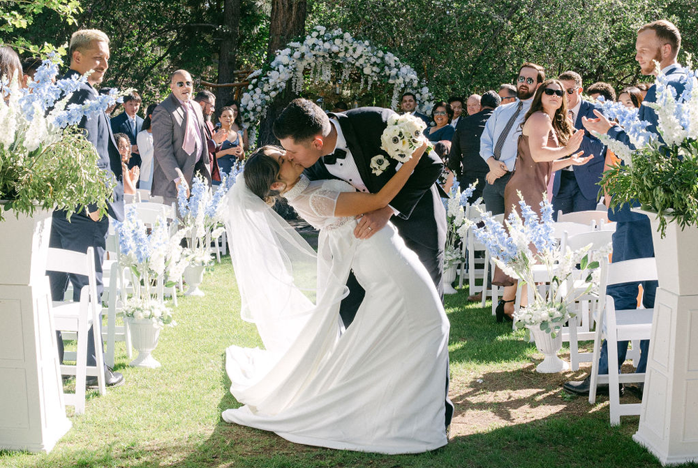 A groom in a tuxedo kisses his bride in a white dress as they stand in the aisle at an outdoor wedding, surrounded by cheering guests, flowers, and greenery—perfectly capturing the romance of Big Bear wedding seasons.