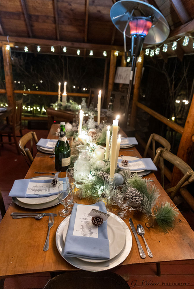 A wooden table set for dinner with blue napkins, candles, plates, wine glasses, a bottle, menus, pinecones, and winter greenery evokes the charm of Big Bear wedding seasons, nestled under a patio heater and string lights in a cozy outdoor setting.